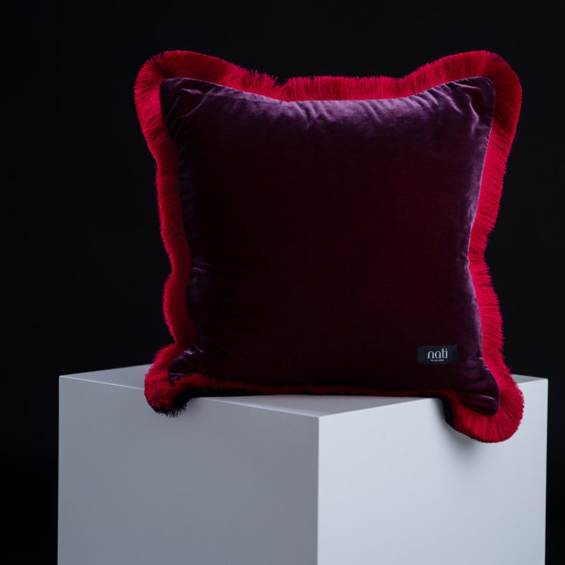 Nati Luxury Pillow Kissen 'Magic – Fleur de grenadier' – auberginefarbener Samt mit rotem Fransenrand, freigestellt auf einem Podest