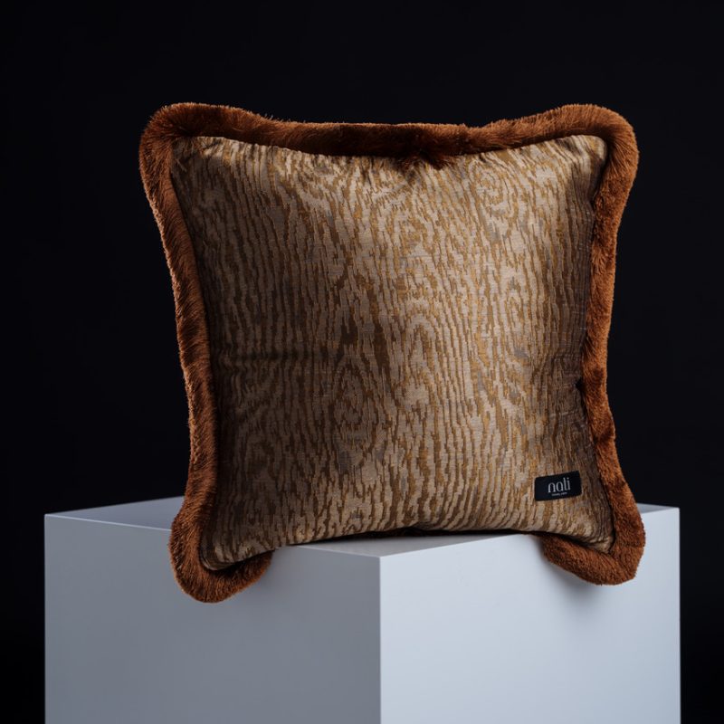 Nati Luxury Pillow Kissen 'Kyoto Bronze' – bronzefarbener Stoff mit Holzmaserungs-Muster und rostbraunem Kunstfellrand, freigestellt auf einem Podest