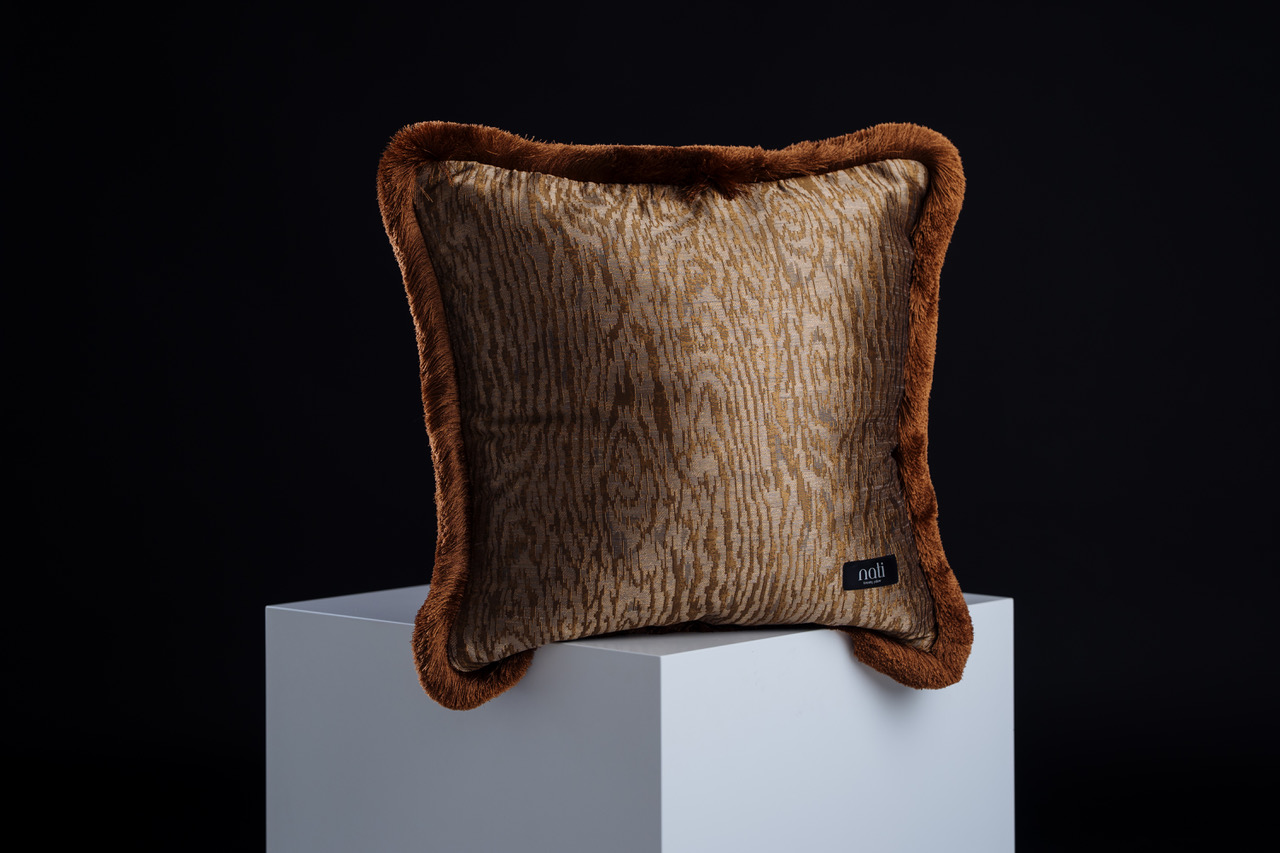 Nati Luxury Pillow Kissen 'Kyoto Bronze' – bronzefarbener Stoff mit Holzmaserungs-Muster und rostbraunem Kunstfellrand, freigestellt auf einem Podest